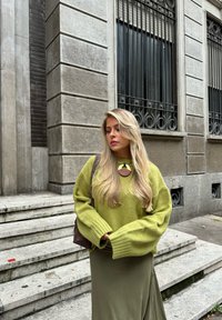 Maglione lavorato a maglia, oversize, di un vivace verde brillante abbinato a una lunga gonna color oliva. Gli accessori includono una collana rotonda marrone. Sullo sfondo ci sono gradini in pietra con texture.