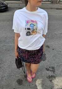 Valge puuvillane T-särk, millel on graafiline print uksest, mis on raamitud lillede ja sõiduki poolt. Kombineeritud musta sädelevate mini-seelikuga ja roosa plätudega.