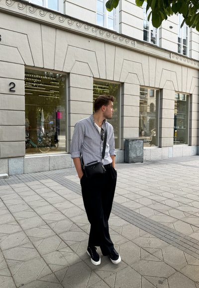 Camisa de botones a rayas, pantalones negros, zapatillas negras con suelas blancas y un pequeño bolso cruzado negro, de pie sobre una acera con patrón.