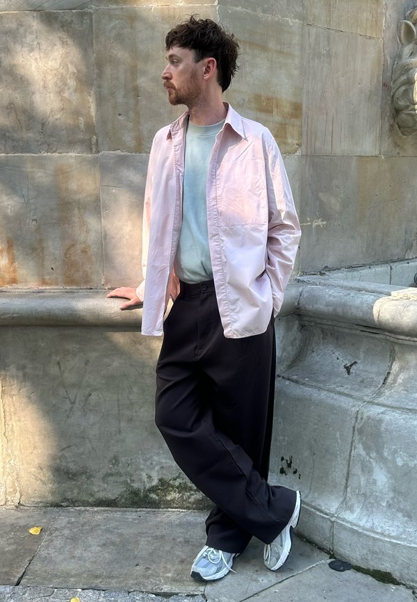 Uomo con camicia rosa chiaro, t-shirt bianca, pantaloni neri e scarpe da ginnastica appoggiato a un muro di pietra che guarda a sinistra in un ambiente urbano esterno.