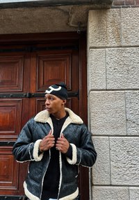 Veste en shearling noire avec doublure et accents en fourrure blanche, portée sur un pull sombre, devant un mur en pierre texturé et une porte en bois en arrière-plan.