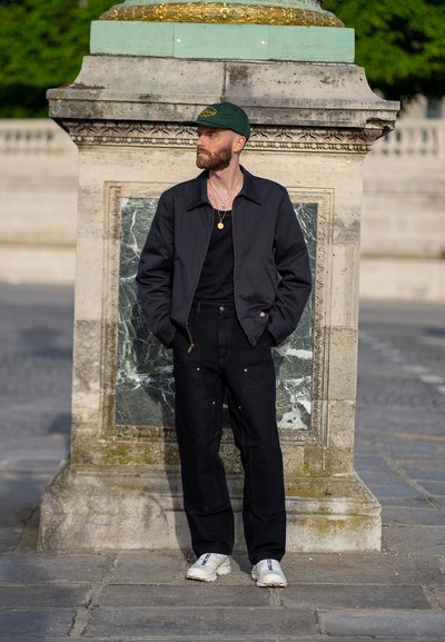 Chaqueta negra sobre una camisa negra, pantalones negros con bolsillos, zapatillas ligeras, gorra verde, de pie junto a un pilar de piedra con acentos verdes.