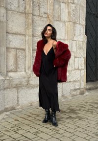 Sort satin natkjole med tynde stropper, kombineret med en dybrød pelsjakke i imiteret pels og sorte ankelstøvler. Står op ad en tekstureret stenvæg.