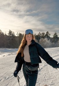 Νεαρή γυναίκα με χειμερινό μπουφάν και γυαλιά σκι κρατώντας μπαστούνια σκι, στέκεται σε χιονισμένη πλαγιά με φόντο δάσος και συννεφιασμένο ουρανό.