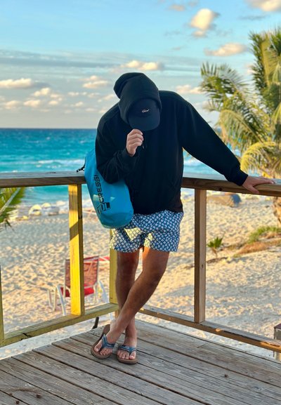 Persona con sudadera con capucha, gorra, pantalones cortos de baño y sandalias apoyada en una barandilla de madera con el océano y la playa arenosa al fondo, sosteniendo una bolsa seca azul.