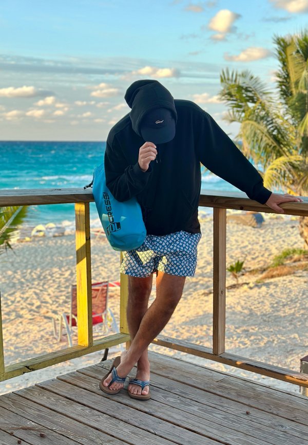 Persona con felpa con cappuccio, berretto, pantaloncini da bagno e sandali appoggiata a una ringhiera di legno con l'oceano e una spiaggia sabbiosa sullo sfondo, che tiene una borsa impermeabile blu.