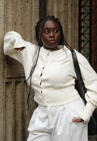 Cardigan blanc à boutons, avec des poignets et un ourlet côtelés, associé à un pantalon blanc taille haute. Le modèle tient un sac fourre-tout noir, s'adossant à un mur.