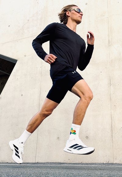 Zapatillas deportivas blancas con rayas negras, pantalones cortos negros y una camiseta de manga larga negra. Corredor con gafas de sol en medio de la zancada contra un fondo de hormigón.