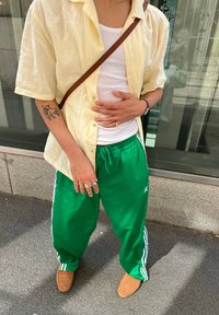Oseba, oblečena v rumeno kratkohlače s kratkimi rokavi, belo majico, zelene Adidas trenerke, svetlo rjave čevlje in rjavo torbico čez ramo, stoji na prostem.