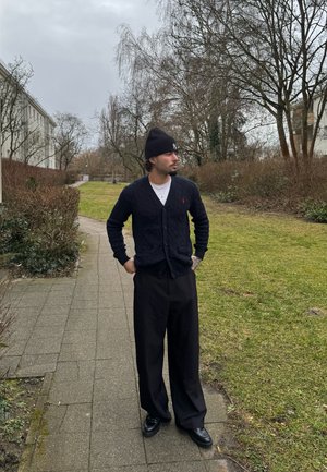 Cárdigan de punto trenzado en color marino con escote en V, pantalones oscuros de pierna ancha y un gorro negro. Zapatos negros, de pie sobre un camino de grava en un parque verde.