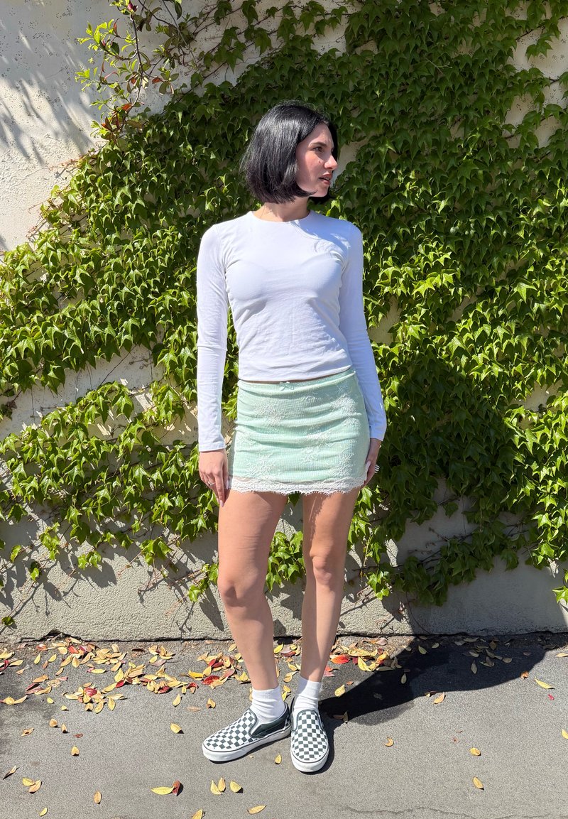 Lühikeste mustade juustega naine, kes kannab valget pika varrukaga topi, mündirohelist seelikut ja ruudulisi tõmbetega kingi, seisab viinamarjaistandusega kaetud seina ääres õues.