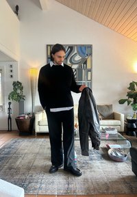 Sort sweater, hvid skjorte med krave, sorte bukser og chunky sorte sko; holder en sort læderjakke i en moderne stue.