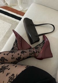 Noge prekrižane, oblečene v črne nogavice z cvetličnim vzorcem in bordo čeveljčke na platformo, poleg temno rjave teksturirane torbice na svetlem kavču.