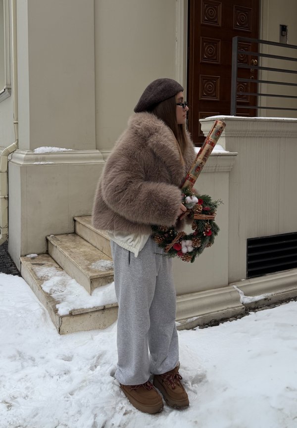 Une personne portant un manteau beige duveteux tient une couronne de Noël décorée et un rouleau de papier cadeau, vêtue d'un pantalon de jogging gris et de bottes marron, dans un décor enneigé.