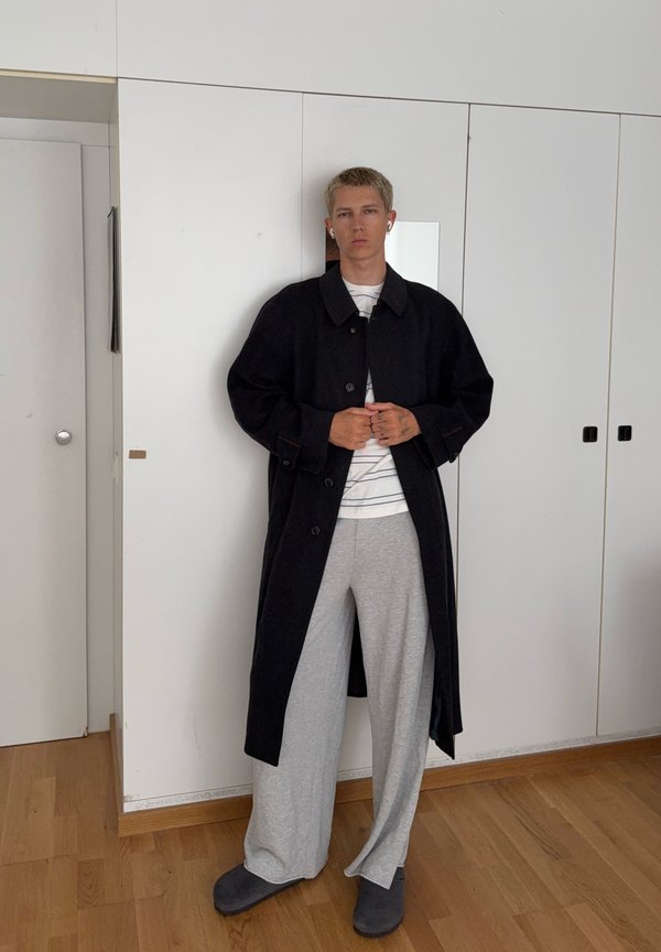 Persona joven con pelo rubio corto viste un abrigo largo negro, pantalones grises anchos, camisa a rayas y zapatillas oscuras, de pie en el interior sobre un suelo de madera.