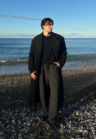 Abrigo largo negro sobre un suéter negro acanalado, combinado con pantalones oscuros y sueltos y botas negras, de pie sobre guijarros junto al mar.