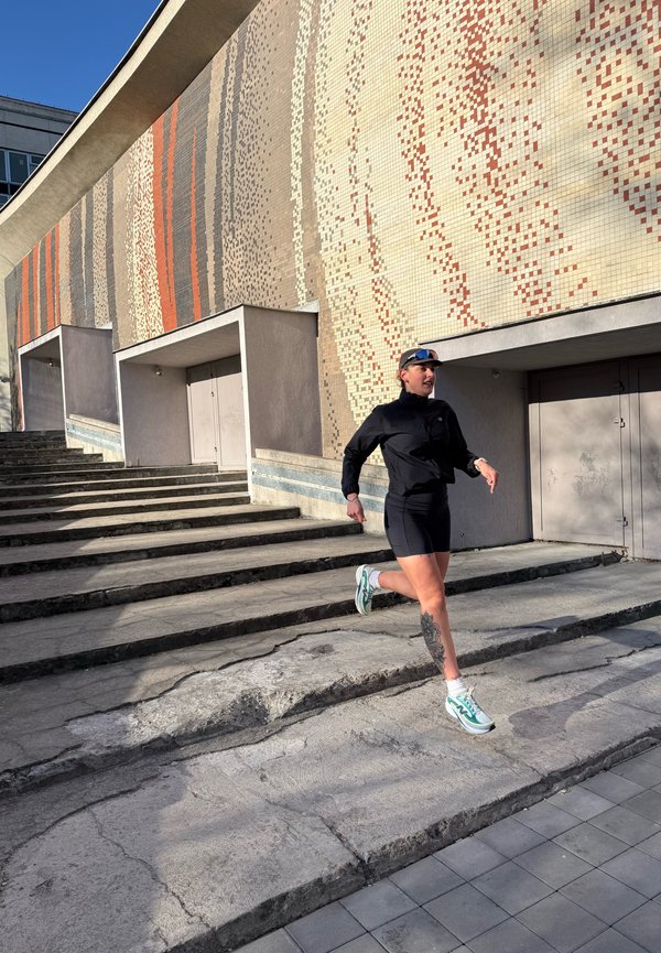 Femme en tenue de sport noire et baskets courant en descendant des marches en béton fissurées à côté d'un grand mur en mosaïque aux motifs orange et beige.
