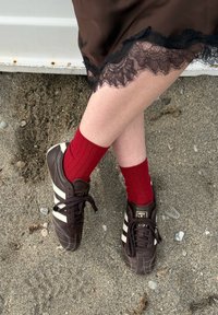 Πόδια σταυρωμένα, φορούν καφέ αθλητικά παπούτσια Adidas με λευκές ρίγες και κόκκινες κάλτσες, μια καφέ φούστα με μαύρη δαντέλα, στέκεται σε χαλίκι.