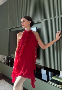 Rotes ärmelloses Kleid mit Rüschen entlang der Säume, hohem Kragen und fließendem Stoff. Das Model posiert drinnen vor einem strukturierten grünen Hintergrund.