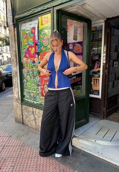 Modelo vistiendo un top de punto azul y pantalones de chándal de pierna ancha negros con rayas blancas. Accesorios de joyería dorada y grandes aretes de aro.