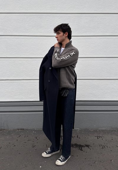 Joven con suéter gris y vaqueros oscuros que sostiene un abrigo largo y oscuro sobre un hombro, de pie frente a una pared blanca con paneles.