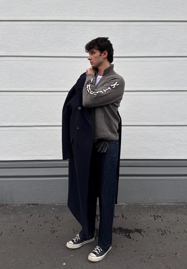 Ung mand iført grå sweater og mørke jeans holder en lang mørk frakke over den ene skulder, stående mod en hvid panelvæg.