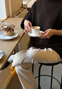 Hvit keramikkopp på et underlag holdt over et trebord. En kaffespiral er synlig. En kanelbolle ligger på en hvit tallerken i nærheten.