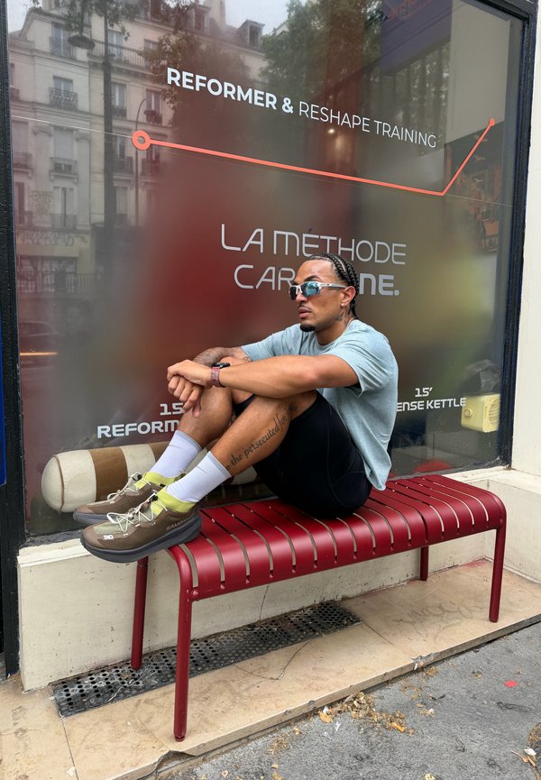 Homme aux cheveux tressés et portant des lunettes de soleil assis en tailleur sur un banc rouge devant la fenêtre d’une salle de sport affichant un texte d’entraînement.