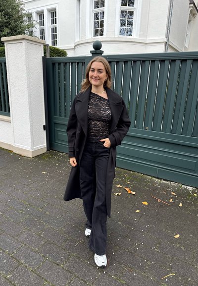 Abrigo largo negro sobre un top de encaje negro, combinado con pantalones negros acampanados y zapatillas blancas, sobre un fondo de una puerta verde.