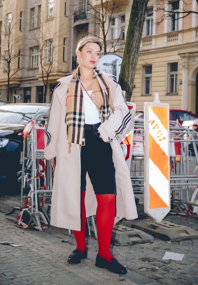 Trench coat beige, camiseta blanca, pantalones cortos negros, medias rojas brillantes y zapatos negros. Presenta una bufanda estampada. Entorno urbano con señales de construcción.