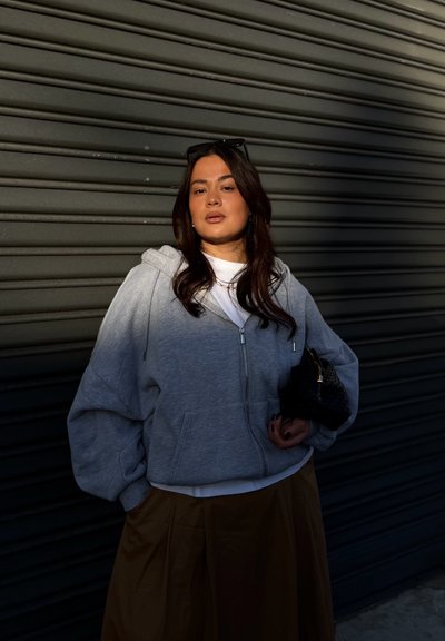 Mujer con cabello largo y oscuro, lleva una sudadera gris con cremallera, camiseta blanca y falda marrón, sosteniendo un bolso negro, de pie frente a una pared de persiana metálica oscura.