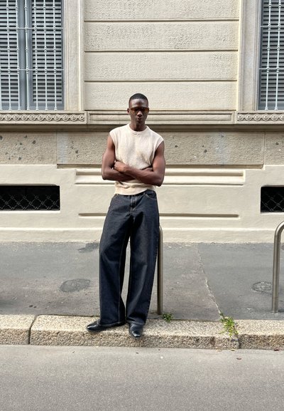 Top de punto sin mangas en color beige, jeans de mezclilla oscuros de pierna ancha y zapatos negros. La modelo está de pie con los brazos cruzados contra una pared texturizada.