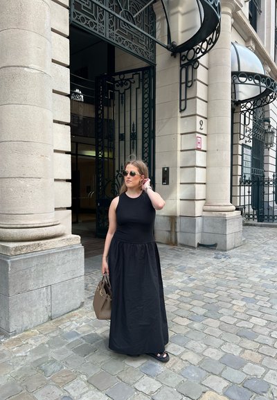 Vestido negro sin mangas con un top ajustado y una falda fluida, combinado con gafas de sol oscuras y un bolso marrón sobre un pavimento de adoquines.