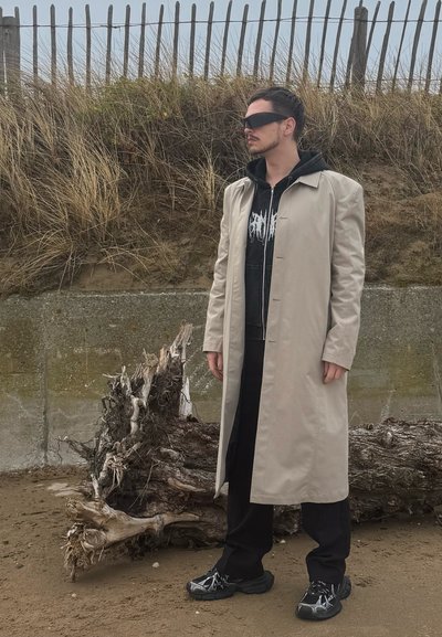 Abrigo largo beige sobre sudadera negra, pantalones negros y zapatillas negras con detalles blancos, de pie junto a un tronco de madera flotante en un suelo arenoso.