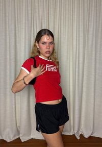 Ung kvinna med långt hår klädd i röd Puma-t-shirt, svarta shorts, håller en svart axelväska, stående framför beige gardiner.