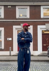 Hombre con gorra roja y chaqueta de mezclilla azul y pantalones del mismo material está de pie con los brazos cruzados en una calle de adoquines frente a un edificio de ladrillo con ribetes blancos.