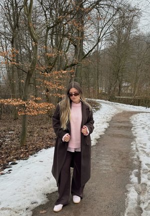 Mujer con abrigo oscuro y suéter rosa caminando por un sendero nevado en un bosque sin hojas durante el invierno.