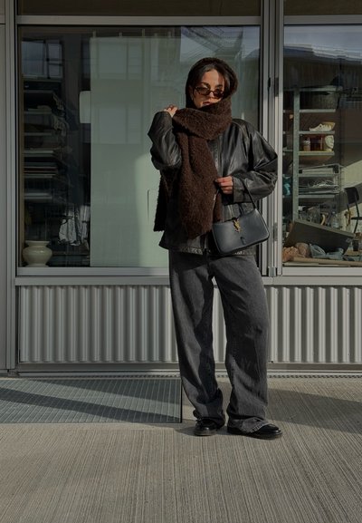 Chaqueta de cuero negra, bufanda texturizada marrón, pantalones gris de corte holgado y bolso negro con herrajes dorados. Las gafas de sol completan el look.