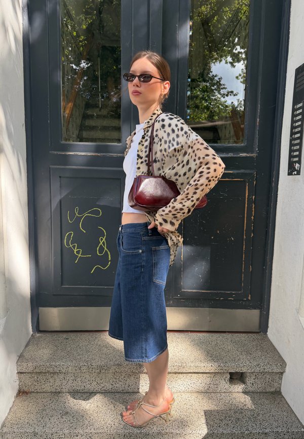 Woman wearing sunglasses, leopard print jacket, white top, denim skirt, and strappy heels standing on outdoor steps in front of a dark door.