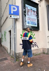 Χρωματιστό ριγέ μπουφάν με κείμενο "JEANS", τζιν σορτς και καφέ μπότες. Αστικό υπόβαθρο με πινακίδα στάθμευσης και αφίσα ταινίας.