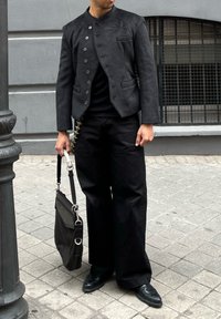 Black suede blazer with a high collar and buttoned front, paired with loose black trousers and a black bag featuring silver hardware.