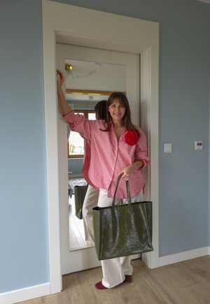 Mujer con camiseta de rayas rosa con detalle de flor roja sosteniendo un gran bolso verde, de pie junto a un espejo de cuerpo entero en una habitación.