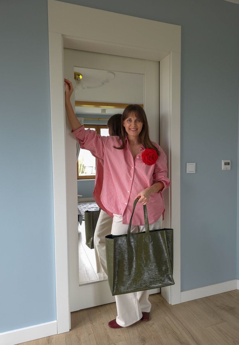 Femme en chemise rayée rose avec détail fleur rouge tenant un grand sac cabas vert, debout près d'un miroir en pied dans une pièce.