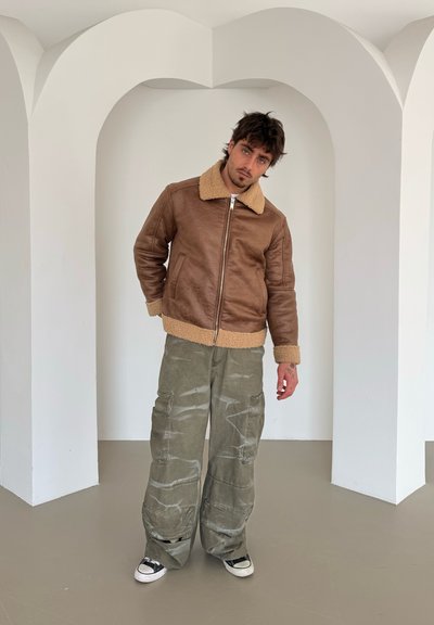 Chaqueta de cuero marrón con un cuello de borreguillo beige, combinada con pantalones cargo holgados de color verde oliva. Unos zapatillas negras y blancas completan el look.