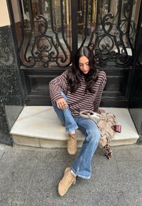 Camicia a maniche lunghe a righe marroni e bianche, jeans a zampa leggeri blu, sneakers beige e una borsa beige con dettagli a catena posata su un gradino di marmo.