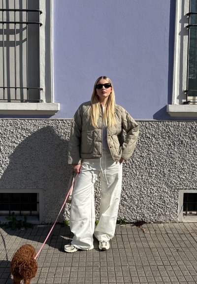 Chaqueta bomber acolchada de color verde oliva, sudadera blanca con capucha y pantalones anchos blancos. Gafas de sol negras y un perro pequeño marrón con una correa rosa. Fondo de pared texturizada.