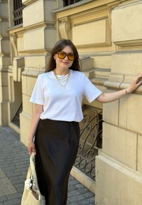 Junge Frau mit gelben Sonnenbrillen, weißem T-Shirt, schwarzem Rock und goldenen Halsketten steht auf dem Bürgersteig einer Stadt und hält eine geflochtene Tasche.
