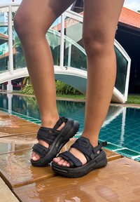 Sandali neri con piattaforma, cinturini regolabili, suola in gomma e finitura testurizzata, indossati su un piede accanto a una piscina su un ponte di legno.