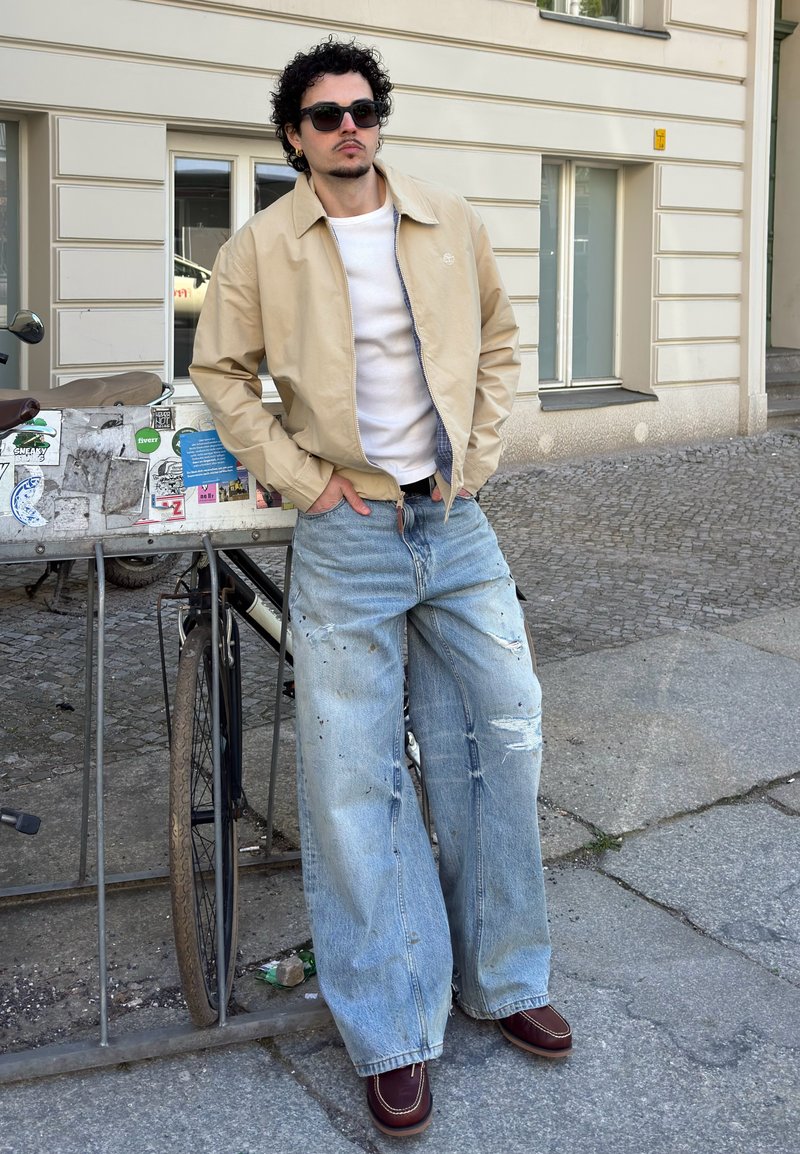 Homme portant une veste beige, une chemise blanche, un jean large déchiré et des lunettes de soleil, appuyé contre un support à vélos sur un trottoir en ville.
