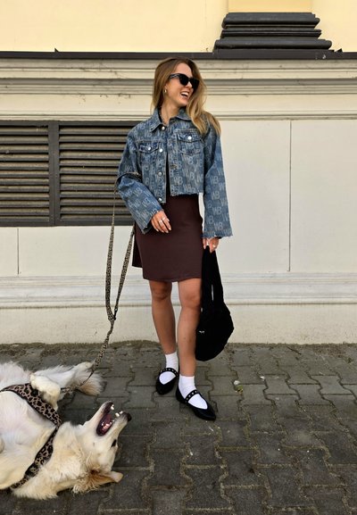 Chaqueta de mezclilla con patrón de logo, vestido marrón, calcetines blancos, zapatos negros con detalles. Un perro yace cerca sobre adoquines.